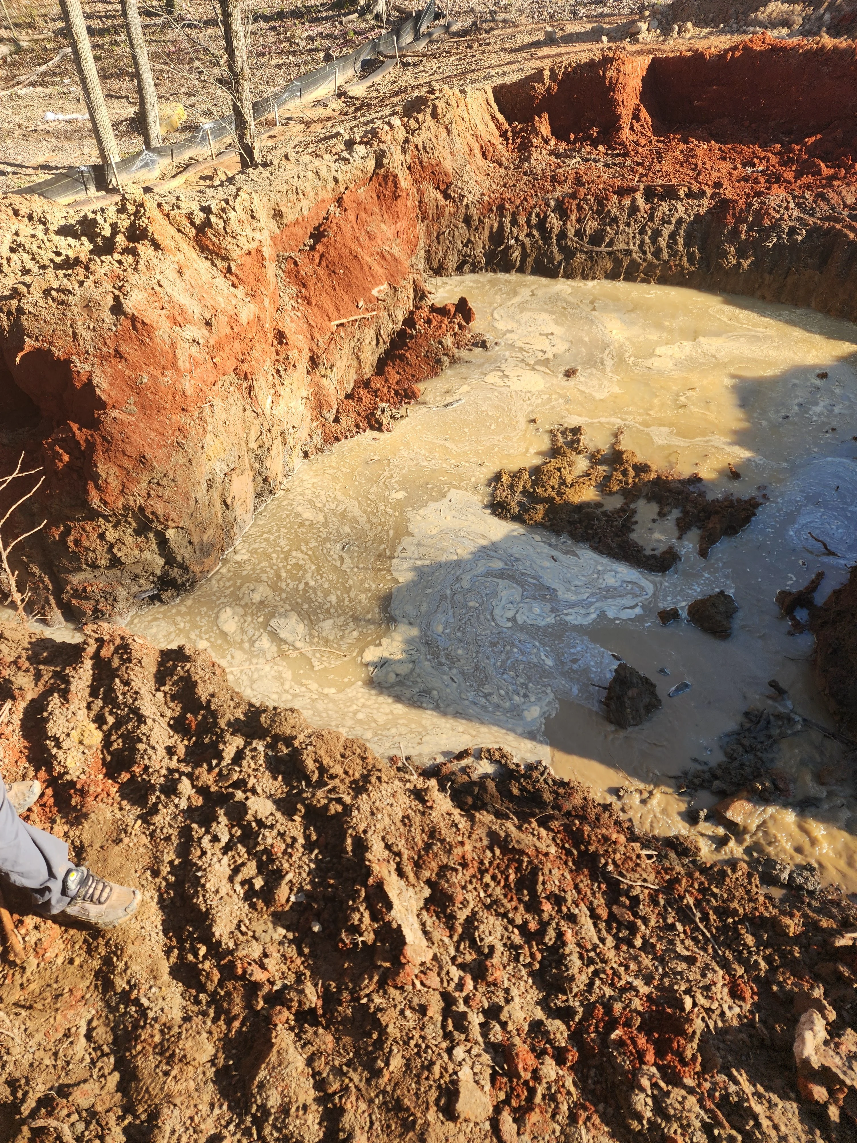 Large excavation pit with red clay walls and water pooling at bottom during foundation work