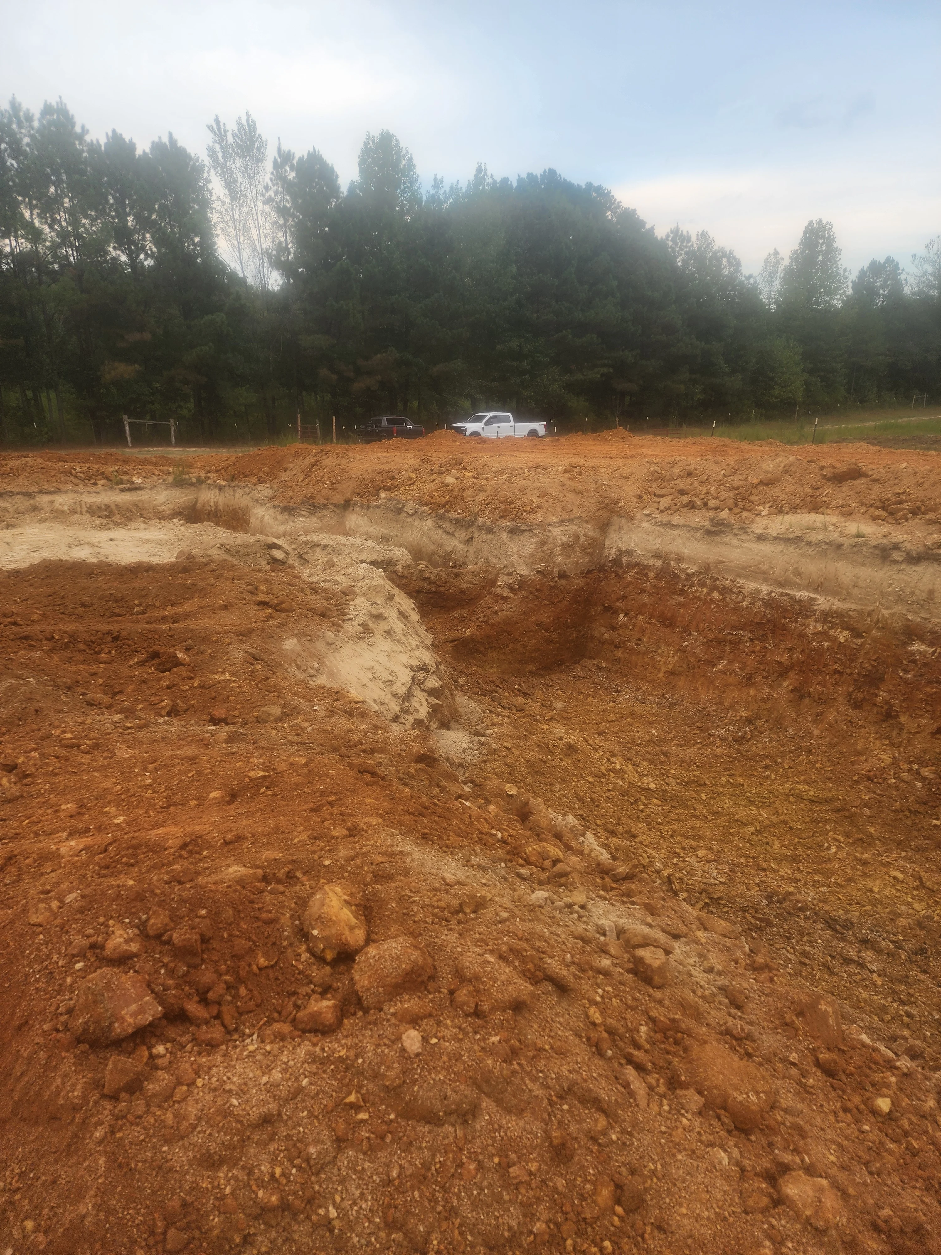 Excavated residential lot with deep trenches and graded earth, forested backdrop