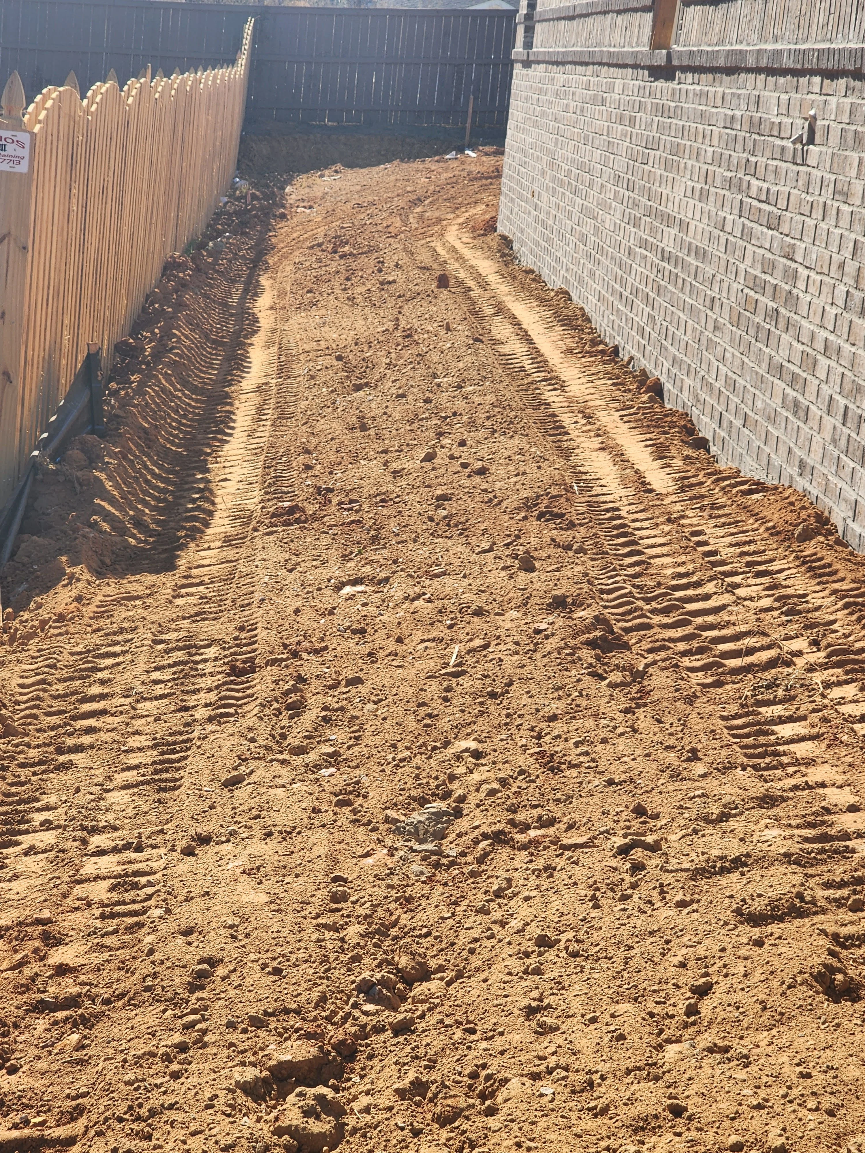 Freshly excavated and graded driveway base with compacted earth between brick wall and metal fence