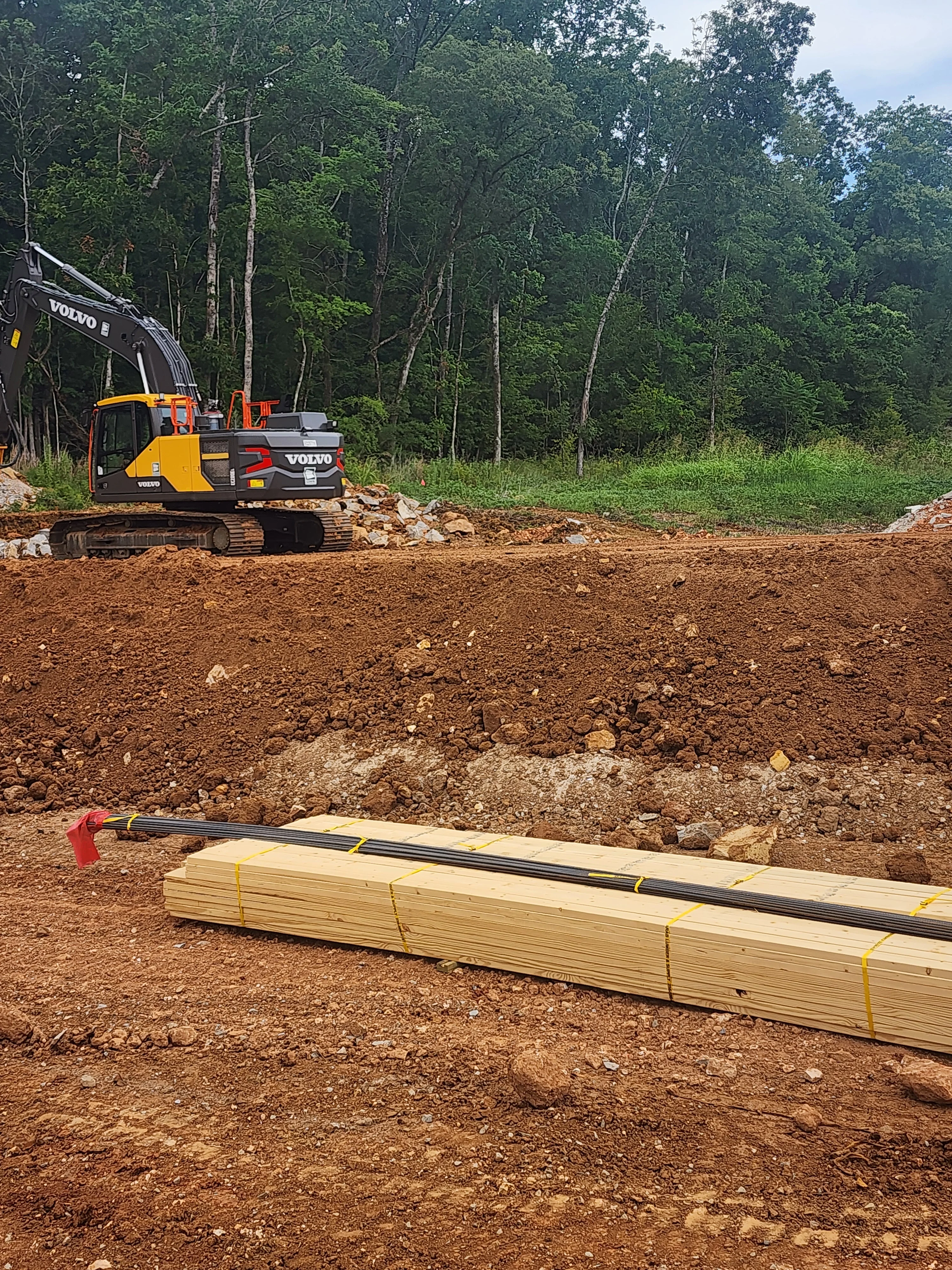 Volvo excavator on cleared dirt site with stacked lumber and forest backdrop in Toney, AL