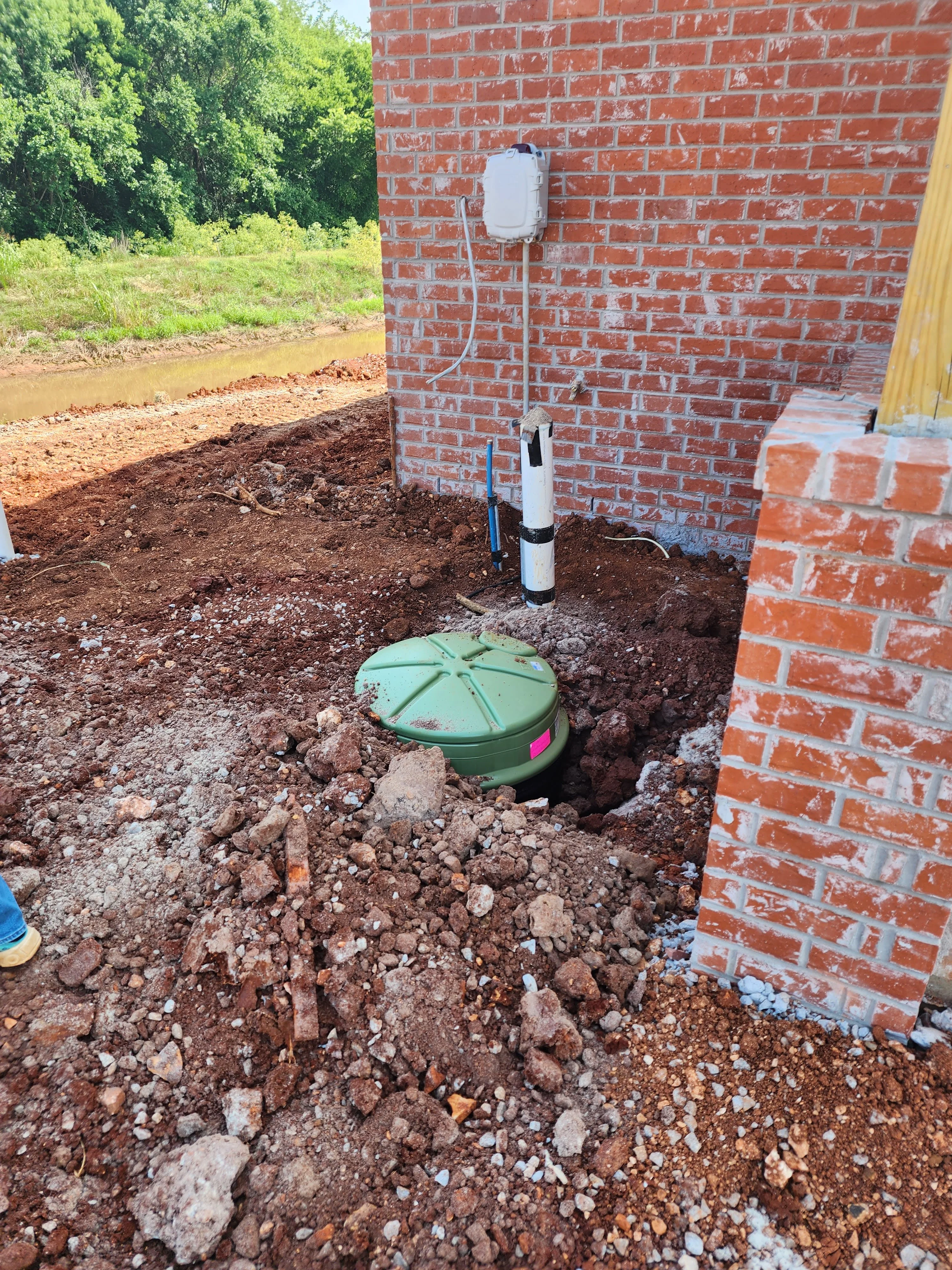 Septic tank installation with green tank, white PVC pipes, and excavated pit beside brick building