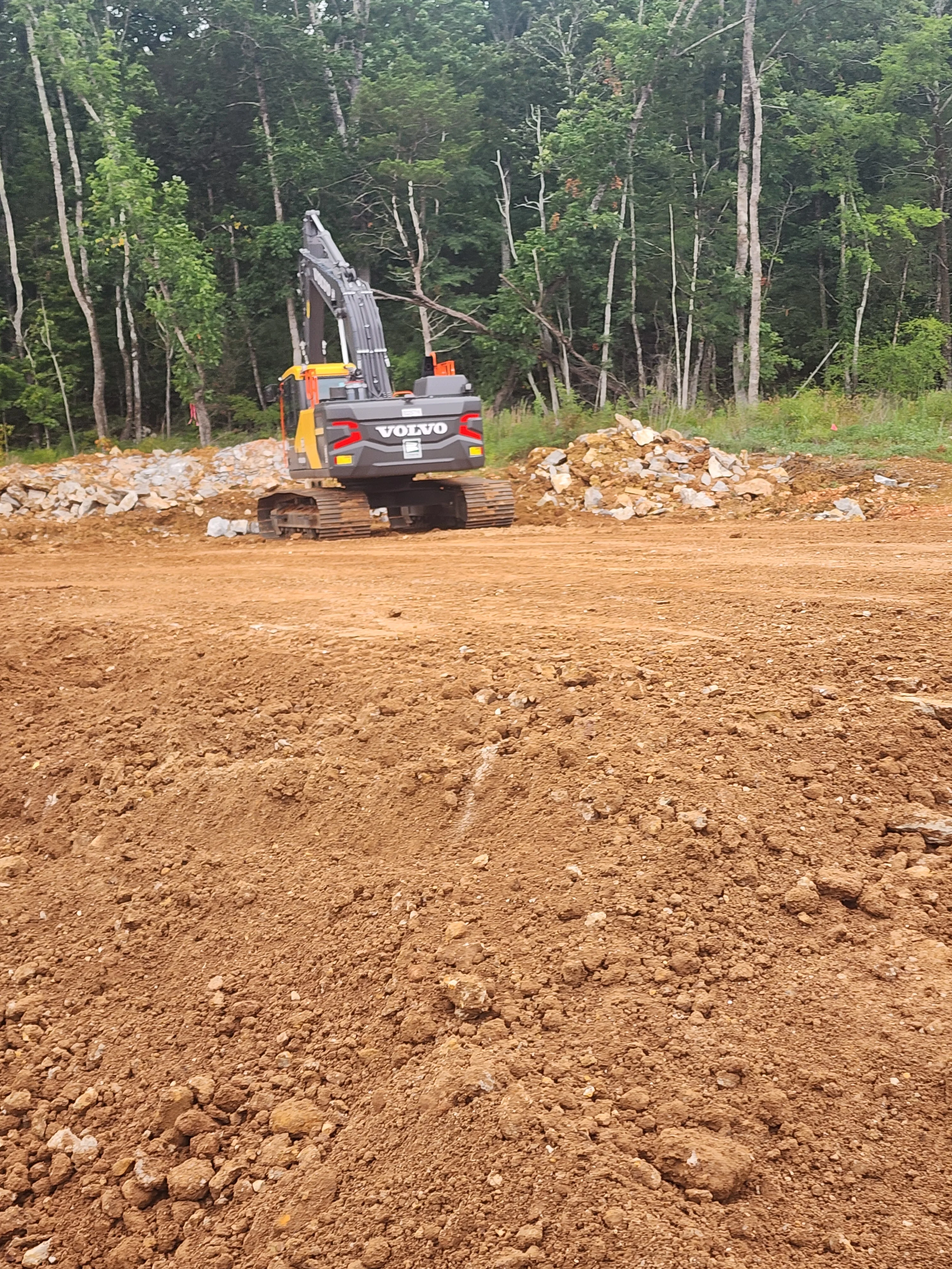 Yellow and black Volvo excavator moving debris on cleared dirt lot with forest backdrop