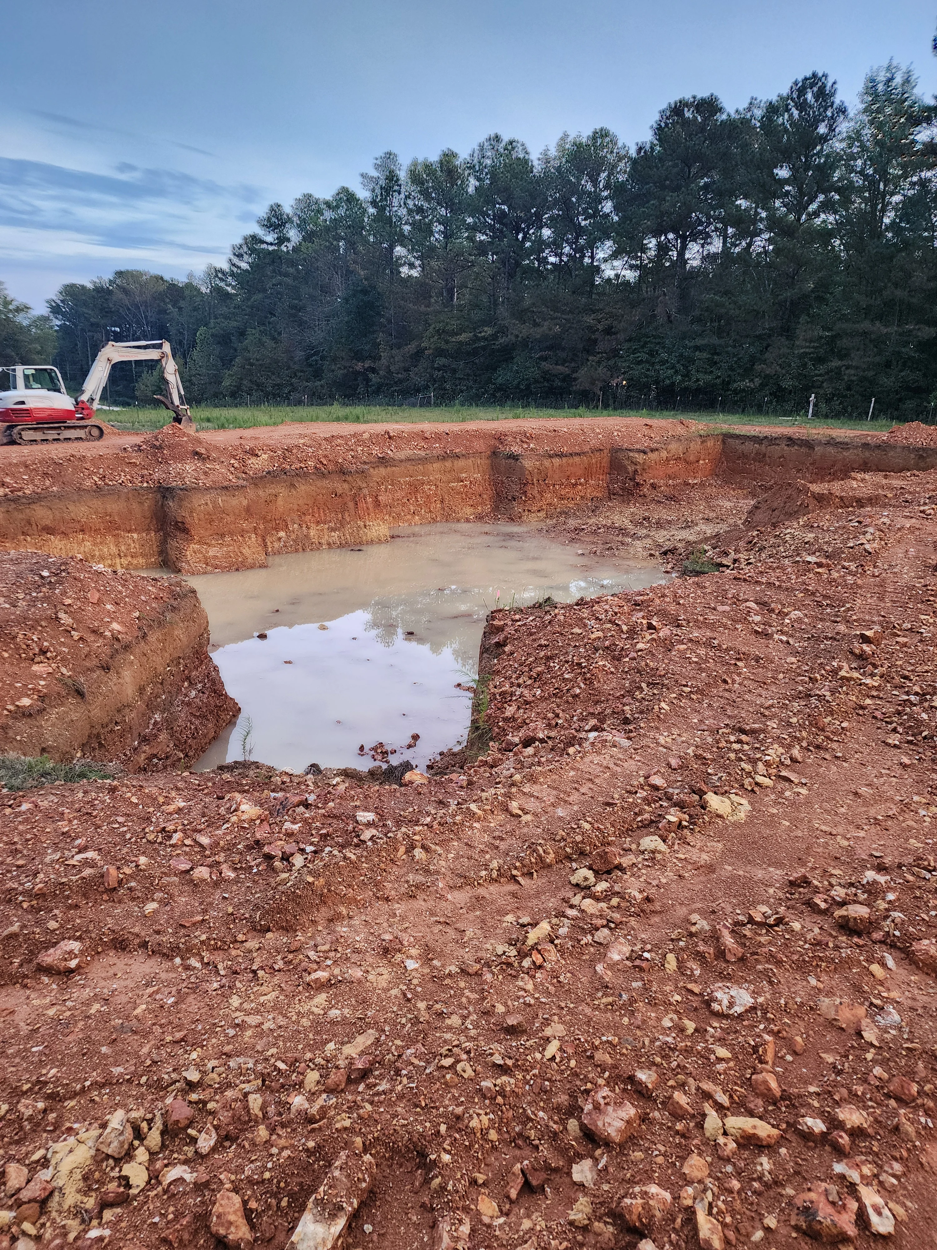 Excavated pond basin with water pooling, red clay banks, and excavator on rural property