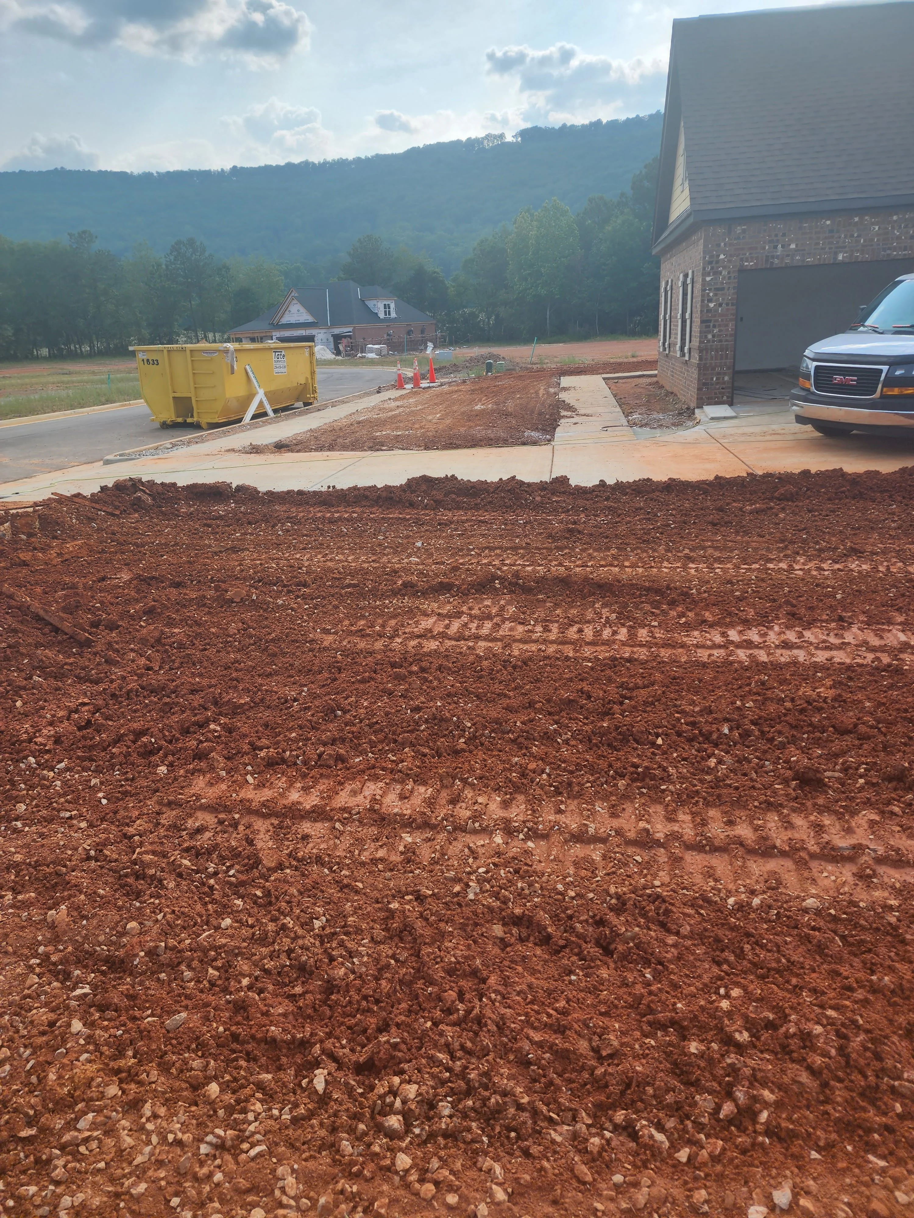 Excavated residential lot with graded red clay, concrete driveway, and brick home under construction