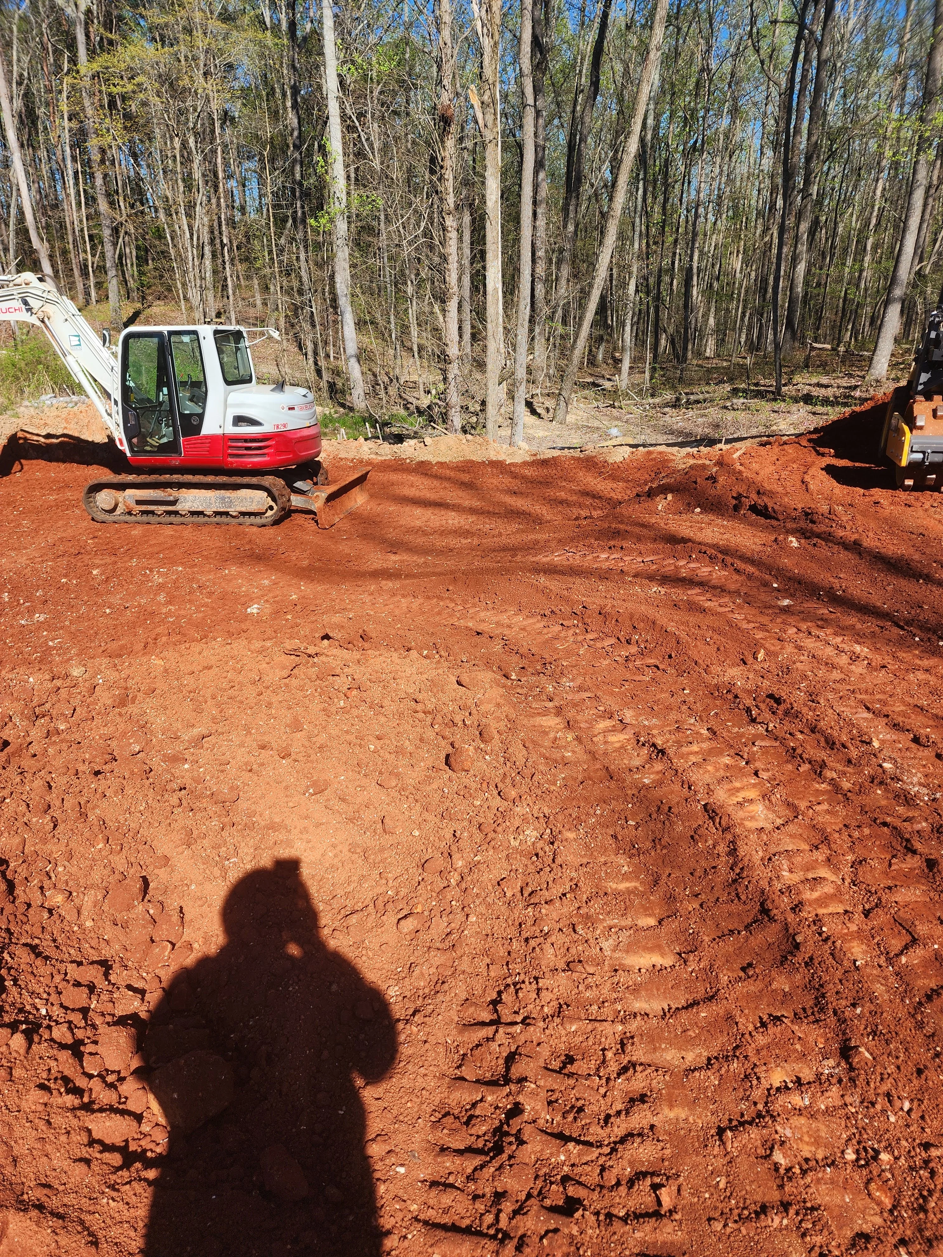 Compact excavator on cleared red clay lot with wooded backdrop