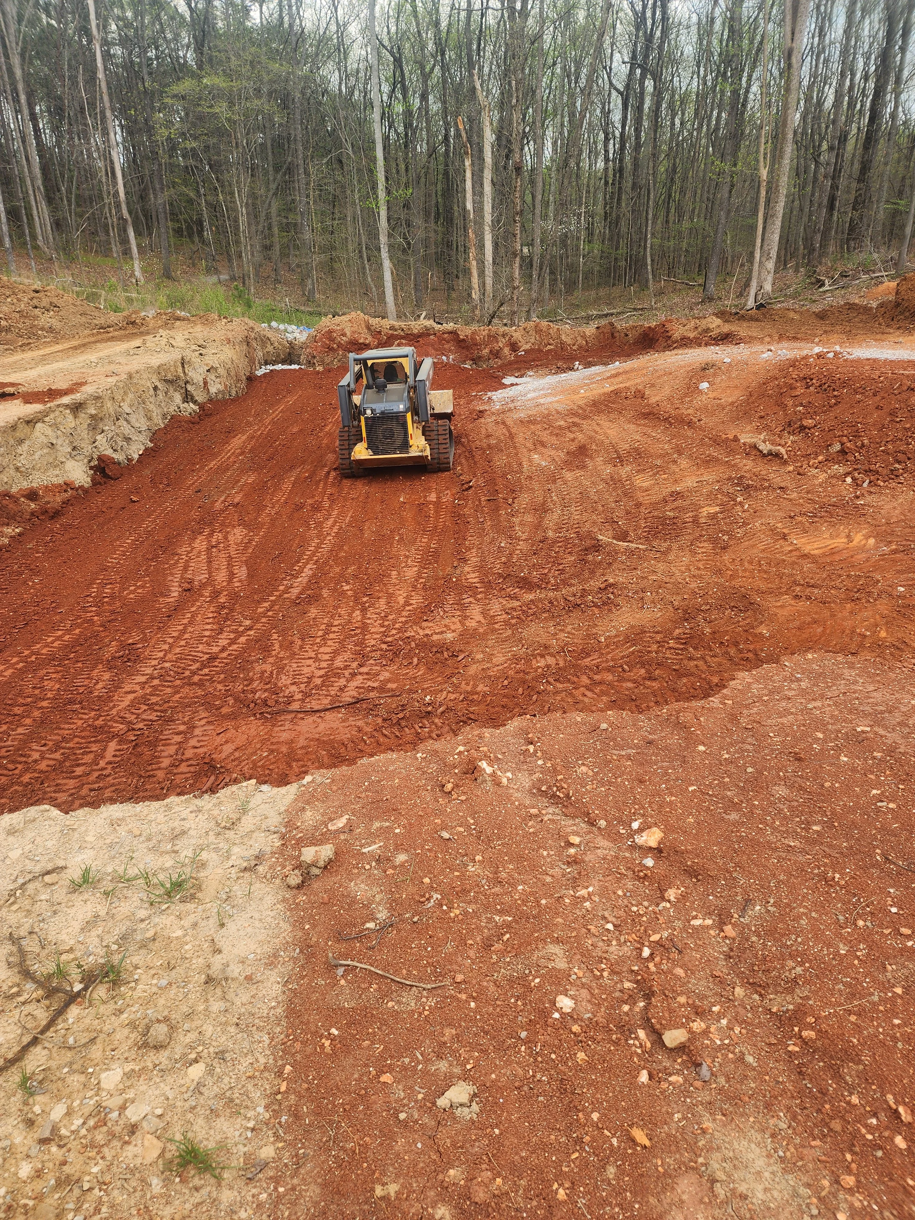 Tracked skid steer grading red clay soil on residential lot surrounded by bare trees