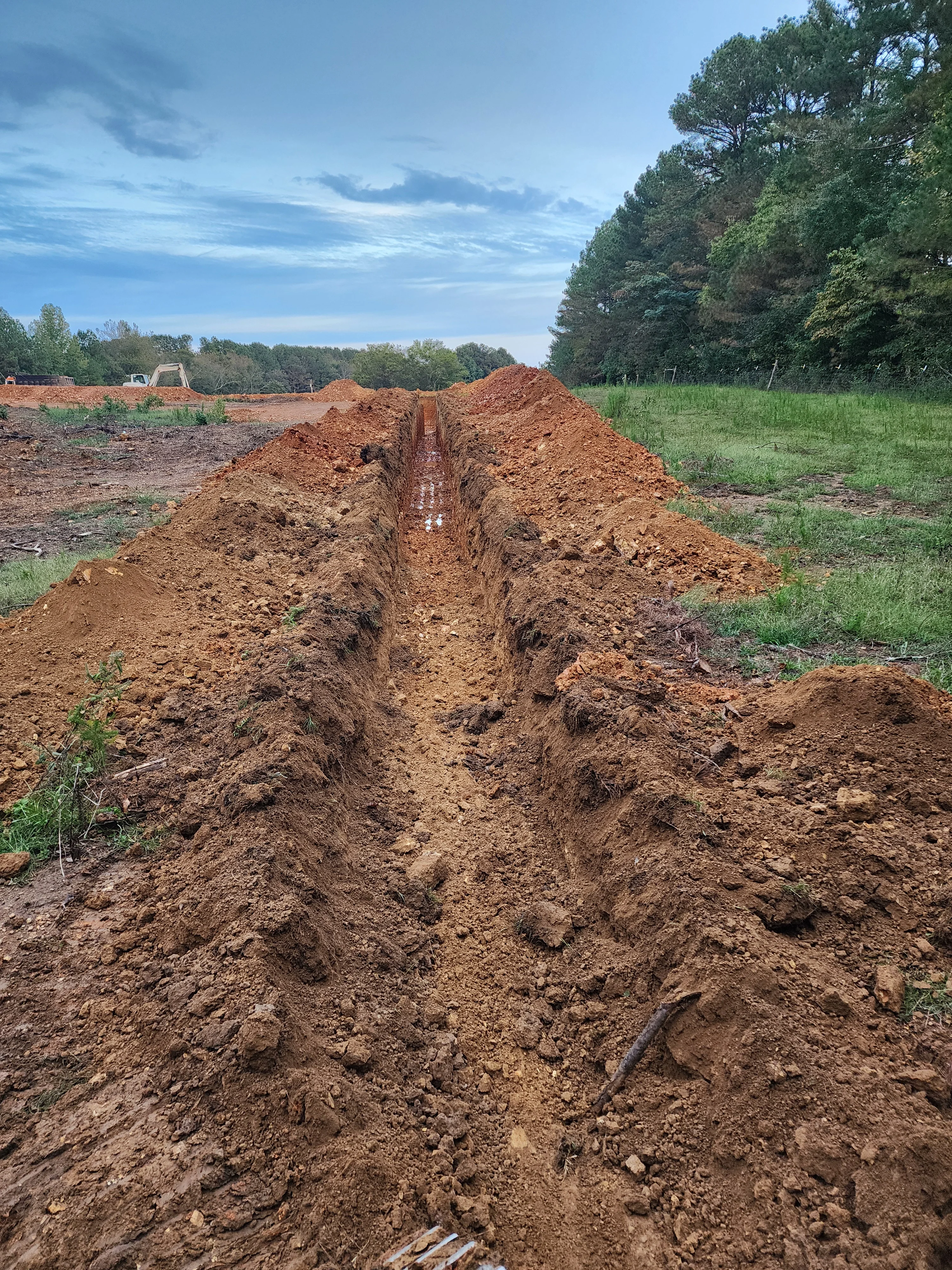 Long excavated trench with clay walls running through rural property with trees and equipment in background