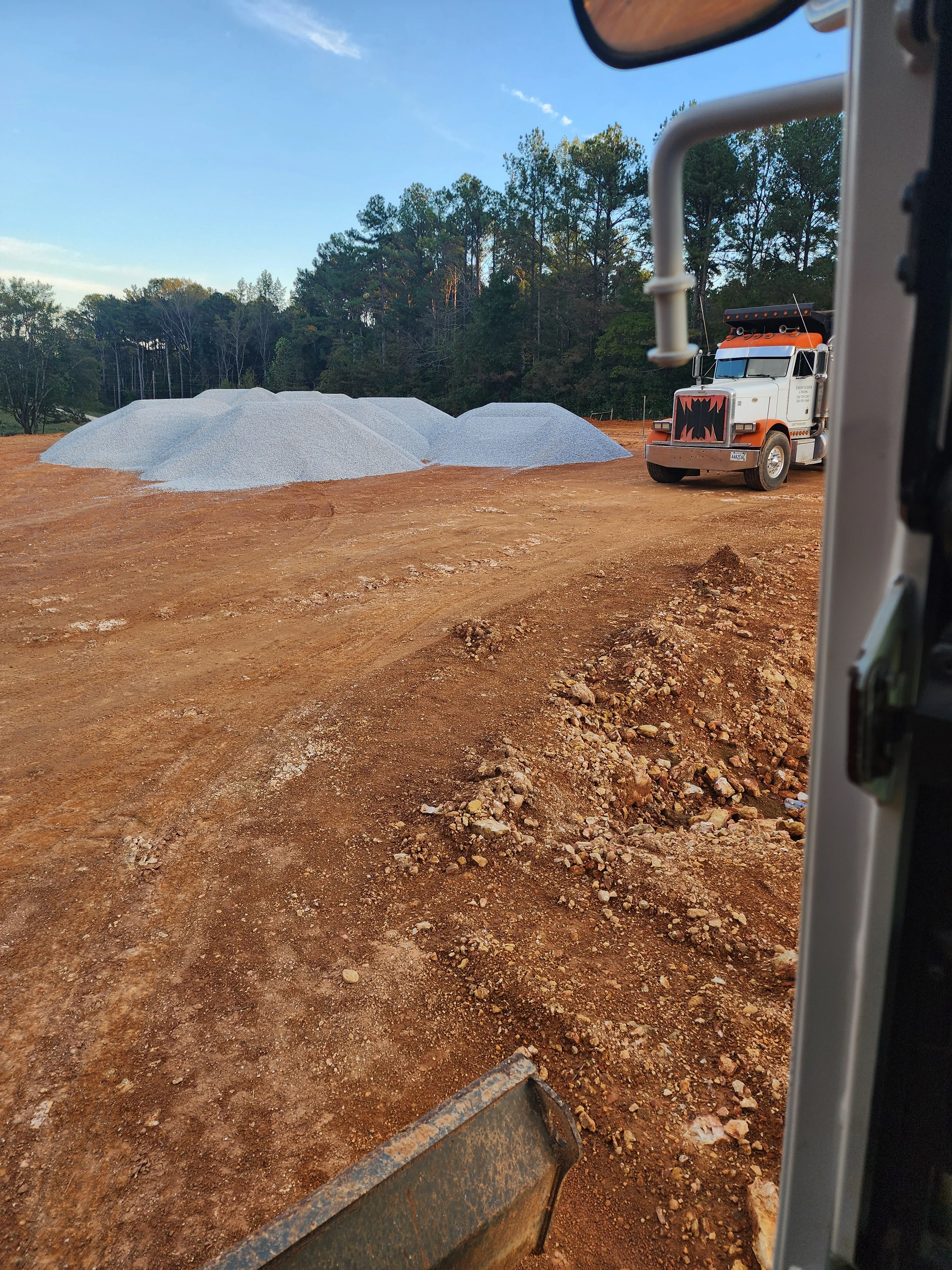 Two large white aggregate stockpiles on cleared red clay site with dump truck and equipment